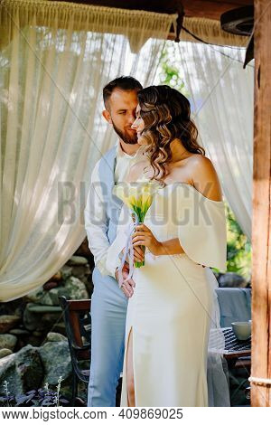 Groom In Blue Suit And Bride With Long Curls With Bouquet In A Gazebo