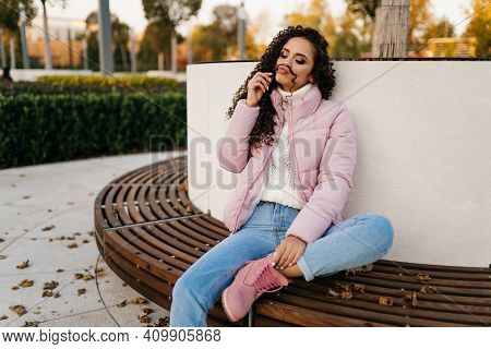 On A Wooden Bench Sits Tucking Under One Leg A Beautiful Girl And Plays The Curl Of Her Long Curly H