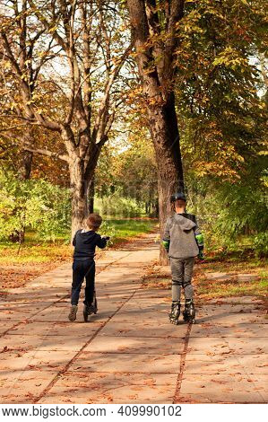 Against The Background Of An Autumn Park, One Boy Is Rollerblading, The Other Is Riding A Scooter.wa