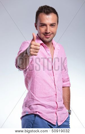 smiling young casual man showing the thumb up gesture while holding a hand in his pocket. on gray background