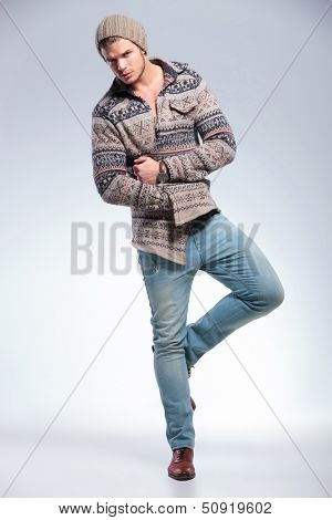 full length picture of casual young man standing on one leg and holding a hand inside his jacket while looking at the camera. on light gray background