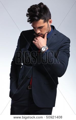 elegant young fashion man in tuxedo looking down while holding his hand on his chin. on gray background