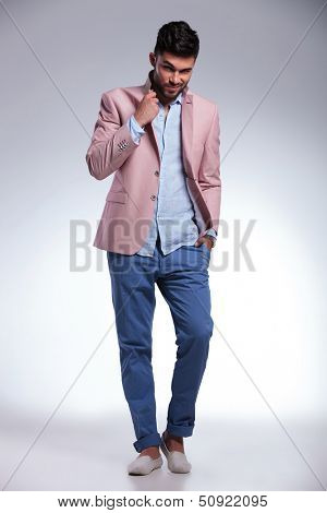 full length photo of a young casual man raising his jacket lapel and holding a hand in his pocket while looking into the camera with an eyebrow raised. on gray background