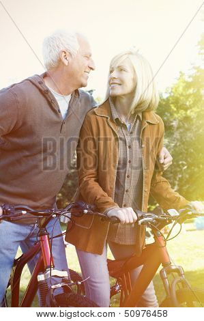 Two aged smiling people with bycicle over park background