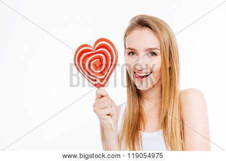 Amusing attractive young woman showing tongue and holding heart shaper lollipop over white background