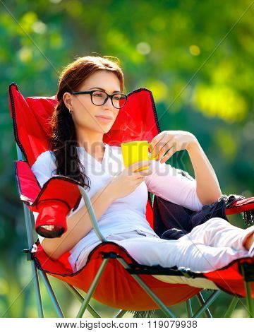 Beautiful woman resting outdoors, sitting on the chair and drinking tea, spending summer vacation in the camp, enjoying calm and peaceful holidays