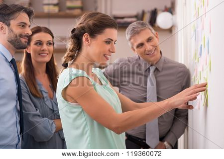 Creative businesswoman pointing sticky notes on wall with colleagues. Group of business people working on new project with many sticky notes. Young creative teamwork working at office. 