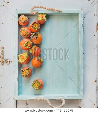 Ripe fresh persimmons on blue wooden tray over light backdrop, top view