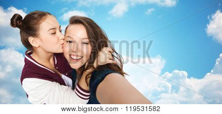 people, friends, teens and friendship concept - happy smiling pretty teenage girls taking selfie and kissing over blue sky and clouds background
