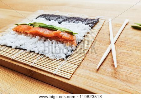 Preparing sushi. Salmon, avocado, rice on seaweed and chopsticks on wooden table. 