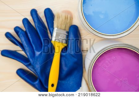 Tin cans of household pink and blue oil paint, a brush and pair of blue latex gloves on a light uncolored wooden background. Close up. Top view. Space for your text or pruduct display.