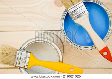 Redecorate a house. Two tin cans of household paint with brushes on top, light uncolored wooden background. Close up. Top view. Space for your text or pruduct display.