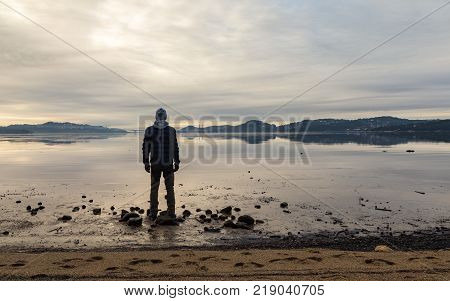 Man standing at the beach in the Norwegian fjord Tovdalsfjorden, looking at the calm sea and the mist and fog. Hamresanden, Kristiansand, Norway