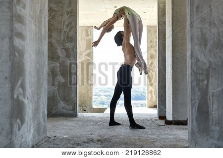 Topless dancer holds a ballerina over his head on the concrete floor of the unfinished building on the cityscape background. Man wears a black dance pants, girl wears a green leotard. Horizontal.