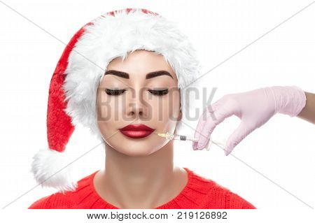 The doctor cosmetologist makes the botulinum injection procedure for tightening and smoothing wrinkles on the face skin of a beautiful young womanin the Santa Claus hat in a beauty salon. New Year's and Cosmetology concept.