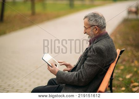 Grandpa use a tablet sitting in the pakr on the bench.