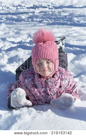 Girl lying on belly in the snow hands in front. The girl narrowed her eyes and wrinkled my nose and smiles. She was pleased and happy. Kid loves the winter holidays.