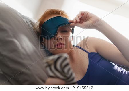 Young woman with sleep mask waking up in bed and snoozing alarm clock on cell phone, mobile telephone. Girl sleeping in bedroom at home and waking up after receiving text message or call on smartphone