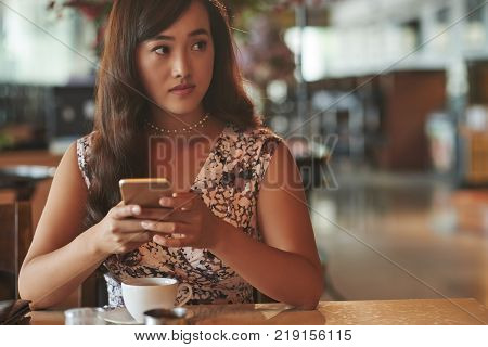 Attractive young Chienese woman having cup of coffee and checking her smartphone