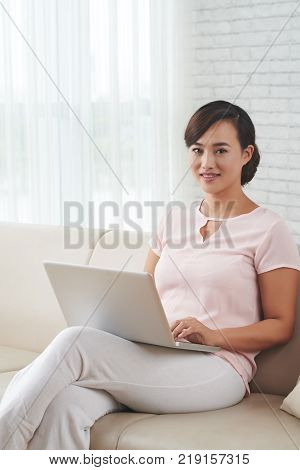 Attractive Chienese young woman sitting on sofa with laptop on her knees