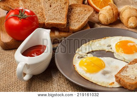 Plate with fried eggs toast with butter croissants bread on wooden cutting board fresh tomato and ketchup in a sauce boat on table covered with sackcloth