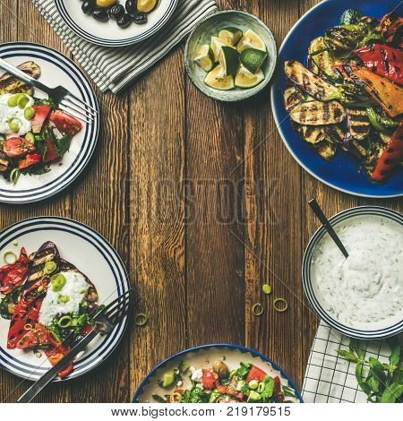 Flat-lay of healthy dinner table setting. Fresh salad, grilled vegetables with yogurt and dill sauce, pickled olives, lemon water over wooden background, copy space, square crop. Clean eating, vegetarian food