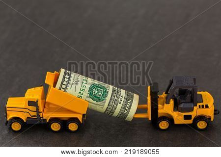 Yellow dump truck carrying dollar money sending to forklift on black stone background. Money transporting via mini vehicles.