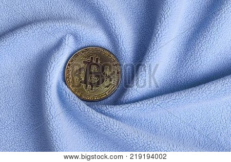 The golden bitcoin lies on a blanket made of soft and fluffy light blue fleece fabric with a large number of relief folds. The shape of the folds resembles a fan from a video card cooler
