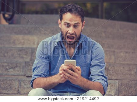 Shocked upset young man looking at his mobile phone seeing bad news or reading text message sitting on stairs outside corporate building. Human emotion reaction expression