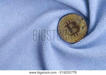 The golden bitcoin lies on a blanket made of soft and fluffy light blue fleece fabric with a large number of relief folds. The shape of the folds resembles a fan from a video card cooler