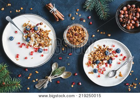 Breakfast composition with homemade cranberry granola, pomegranate, yogurt, spices, nuts and tied tea spoons. Decorated with fir tree branches. Top view