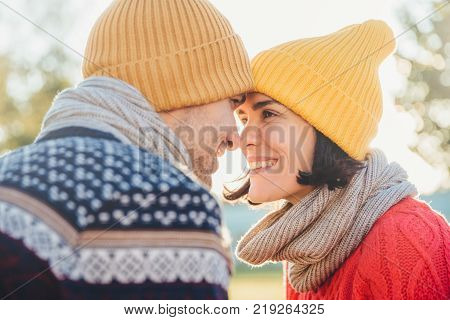 Adorable brunette female and her boyfriend stand closely to each other look at eyes smile happily as feel love and support. Good looking woman with scarf and hat has date with young attractive man