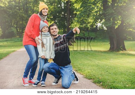 Pleasant looking dad shows with fore finger beautiful landscape to her daughter embraces her. Family of three members have walk at park have good relatioship look into distance as notice something