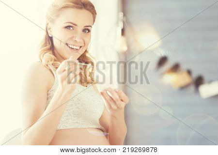 Healthy habit. Cheerful beautiful pregnant woman eating joghurt and smiling while resting at home