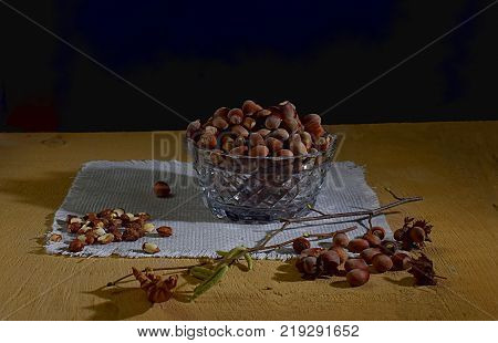 On a sheet of plywood on a napkin there is a crystal salad bowl filled with walnut nuts rows of nut kernels and also nuts and a twig with the flowers of hazel.