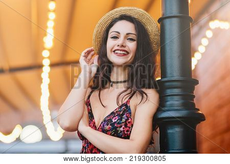 Excited female with dark hair shinig eyes and red lips dressed in dress and summer hat posing at camera with happy and confident expression. Attractive female brunette with make-up resting at cafe