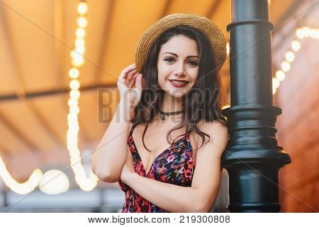 Carefree slim woman with dark luxurious hair shinig eyes and red rouged lips dressed in straw hat and dress while posing at cafe or restaurant. Happy young female having rest at outdoor cafe