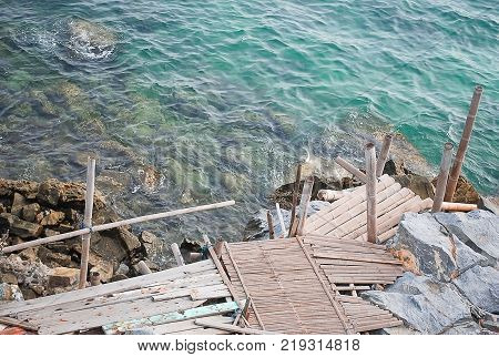 Sidewalk in wood and bamboo at shore close to big stone with backgroung clear blue green the sea and small wave hit the rock.