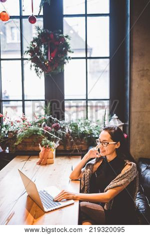 Beautiful Young Brunette Woman Inside Cafe With Christmas Interior At Wooden Table By The Window Tal