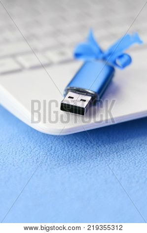 Brilliant blue usb flash memory card with a blue bow lies on a blanket of soft and furry light blue fleece fabric beside to a white laptop. Classic female gift design for a memory card