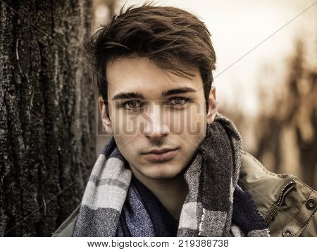 Handsome young man outdoor in winter fashion, wearing dark jacket and woolen scarf in city park looking at camera