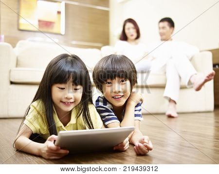 two asian children lying on floor playing video game using digital tablet while parents watching in the background.