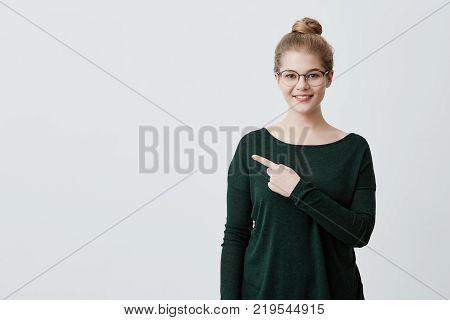 Isolated studio shot of cheerful young female customer or student with blonde hair in casual green sweater and eyeglasses smiling broadly, pointing her index finger at gray copyspace wall for your text or advertising information