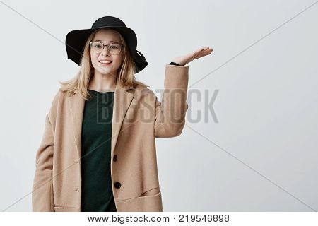 Portrait of young female feeling tired and unhappy, gesturing with open palms, looking up, fed up with stressful bad working day, wants to go out but is afraid of rain. Female student in coat and hat puzzled because of bad weather conditions.