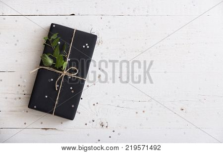 Top view on nice Christmas, birthday gift wrapped in black paper and decorated with cord and tree brunch on white wooden background wuth sillver stars confetti