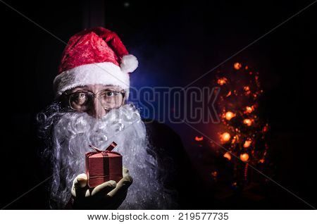 Santa Claus holding gift with festive holiday background. Santa Claus holding and offering a gift box on his hand. Selective focus. Dark toned background