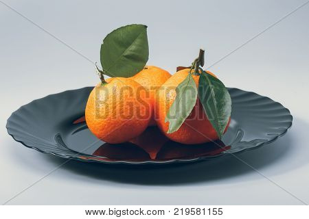 three ripe orange  tangerines with green leaves on a black dish on a white background