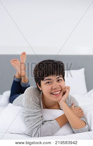 Excited Asian lady enjoying morning in bed and smiling at camera. Jolly young woman waving legs while lying on comfortable bed. Rest concept