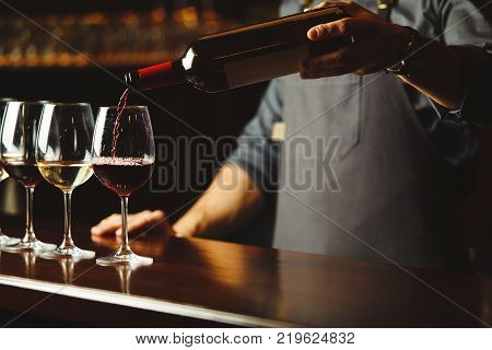 Bartender in apron pours sweet red wine from bottle in capacious glasses on wooden bar counter. Delicious alcohol drink in transparent glassware with nice scent.