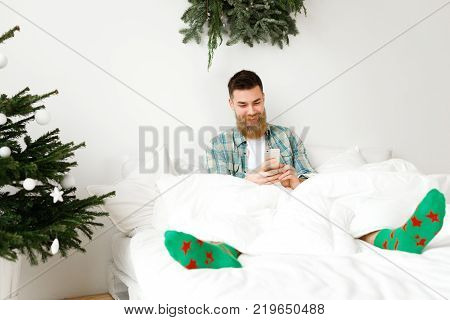 Young cheerful male with long thick beard, wears colorful socks, sits in bed and uses smart phone, being glad to recieve messages of conrgratulation with New Year from friends. Decorated bedroom